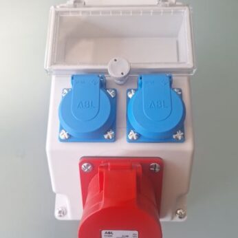 Boîtier de prises électriques industrielles avec une prise triphasée rouge et deux prises bleues protégées par clapets, monté dans un coffret étanche blanc avec capot transparent.