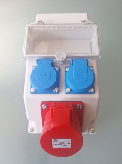 Boîtier de prises électriques industrielles avec une prise triphasée rouge et deux prises bleues protégées par clapets, monté dans un coffret étanche blanc avec capot transparent.
